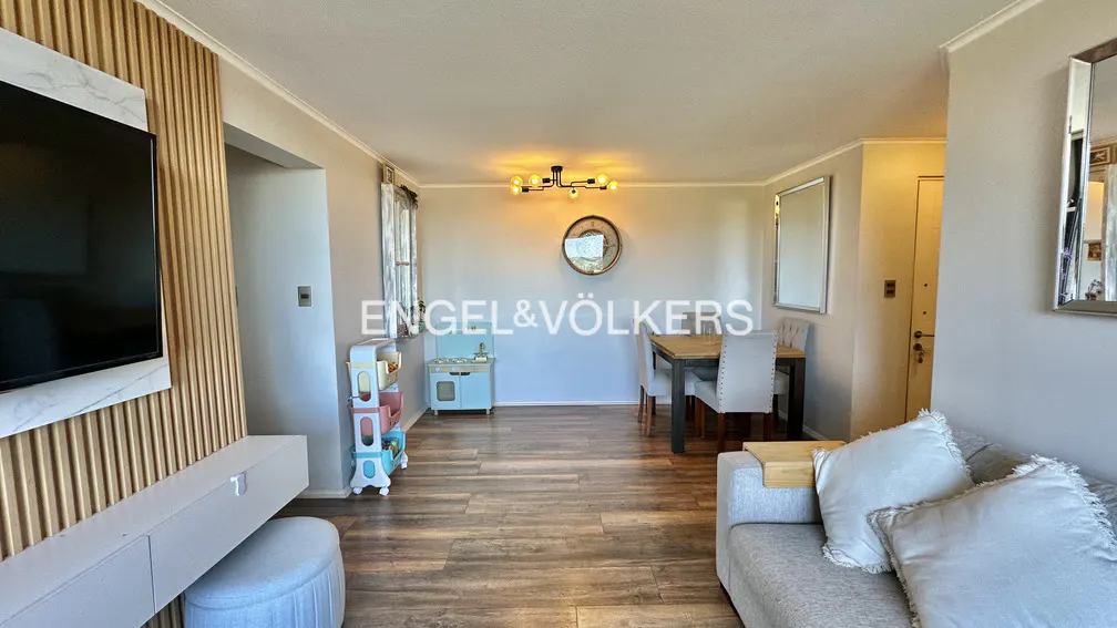 A wide shot of a living and dining area with wood floors, a gray sofa, and a dining table with four chairs. A TV is mounted on a wood-paneled wall. There is a child's play kitchen in the corner.