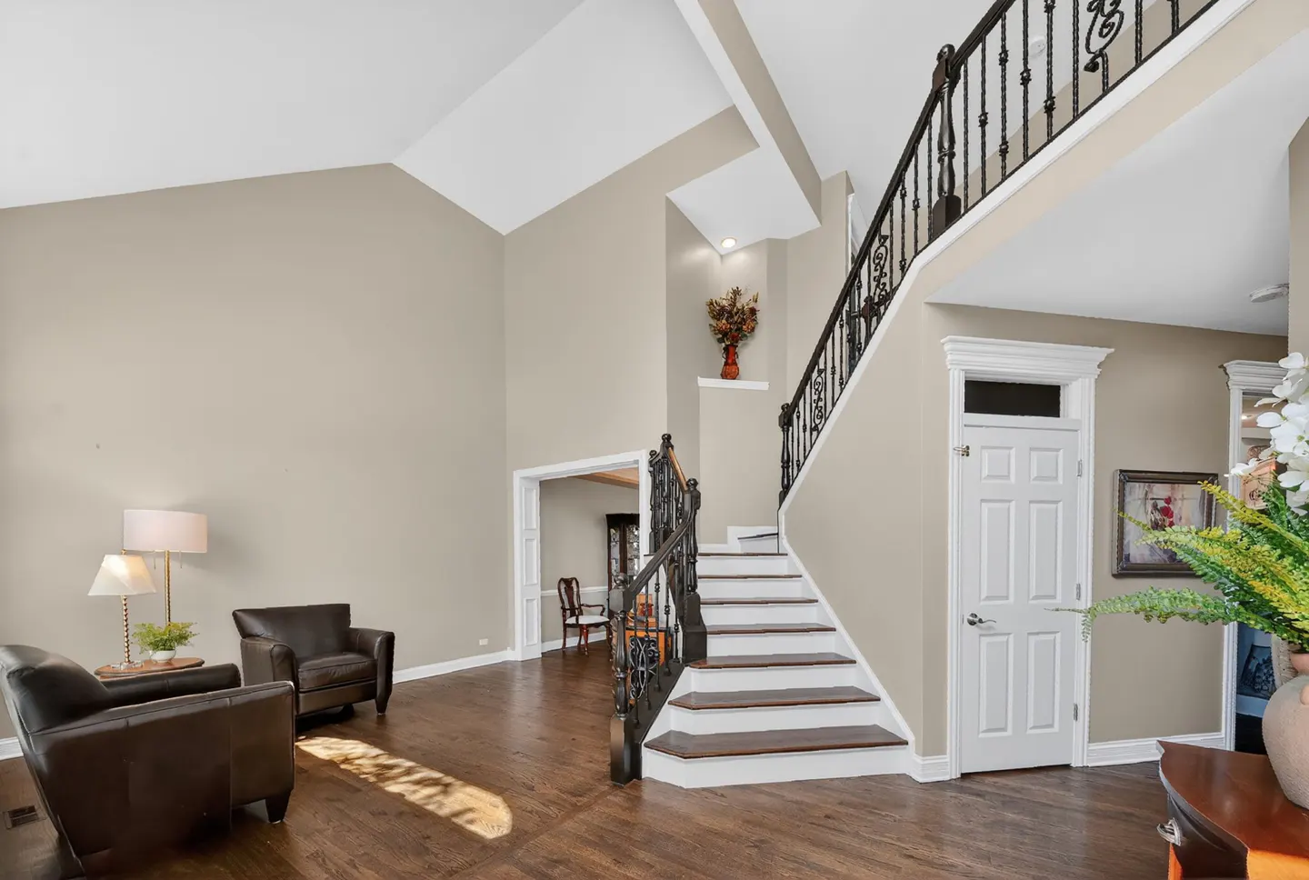 Interior view of a foyer with dark wood floors, beige walls, and a staircase with black wrought iron railing. Two brown leather chairs are on the left.
