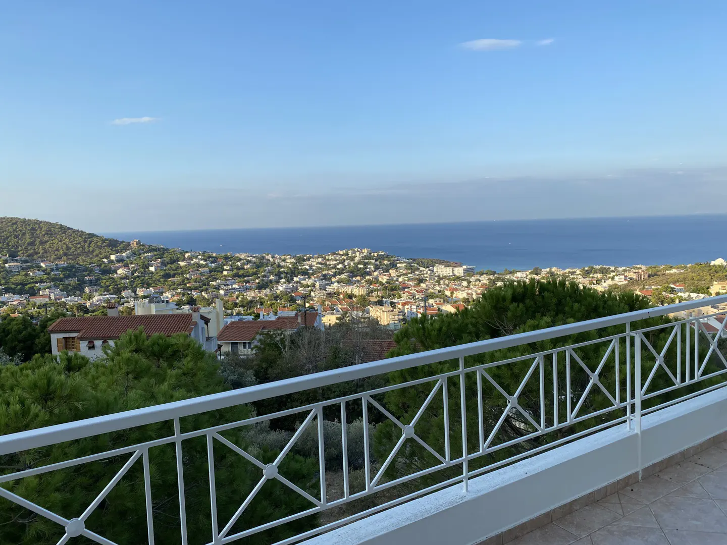 Balcony view of a coastal town. White railing in the foreground, houses with red roofs and green trees below, and the blue sea and sky in the background.