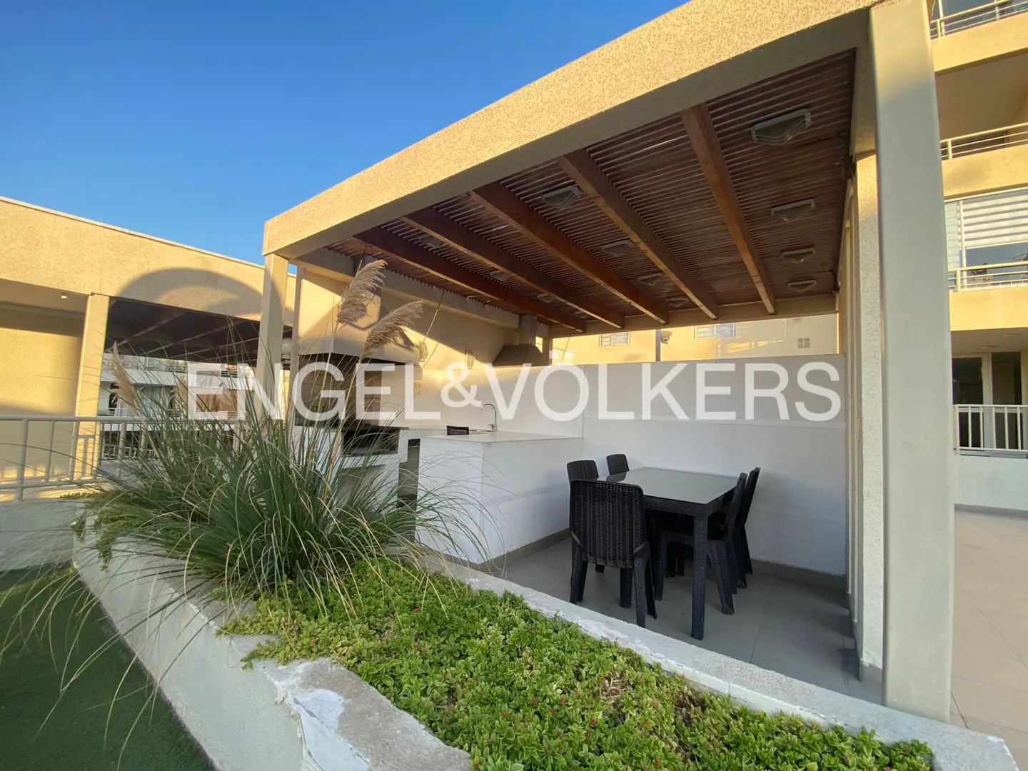 Outdoor dining area with a black table and chairs under a wooden pergola, with lush greenery in the foreground. Engel & Volkers logo visible.