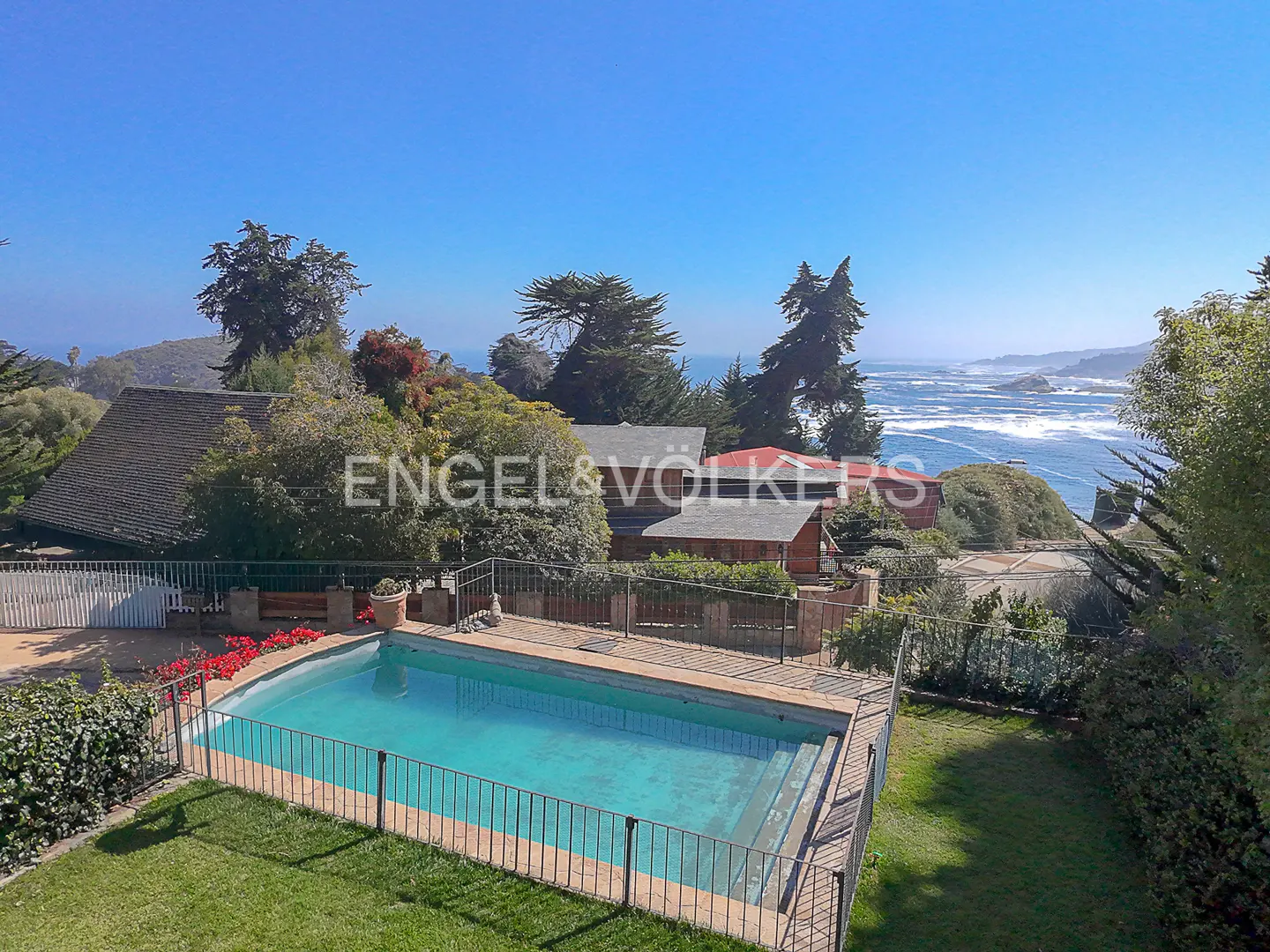 A backyard pool with ocean views. The pool is surrounded by a fence and green grass. The ocean is in the background.