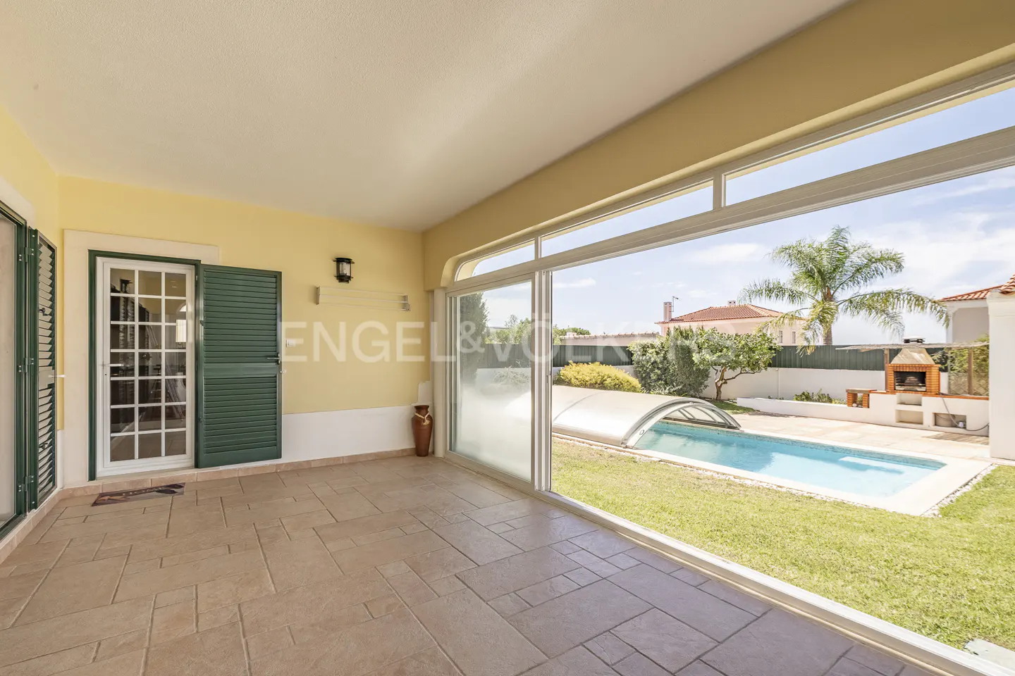 Covered patio with tiled floor and green shutters, overlooking a backyard with a swimming pool and outdoor brick grill.