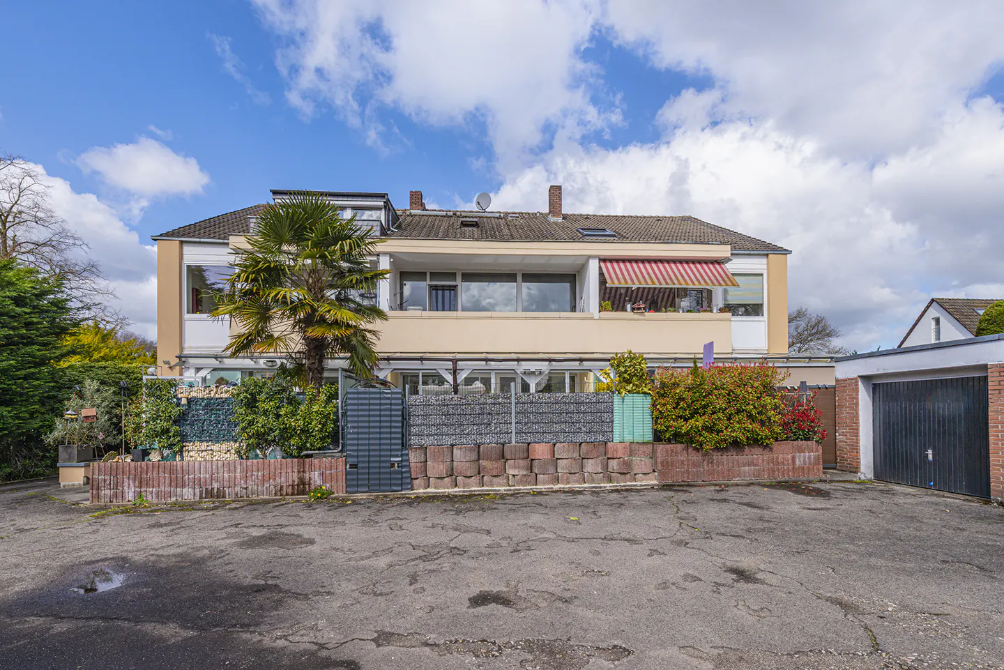 Exterior view of a two-story beige building with a red and white striped awning, a palm tree, and a brick and stone fence.
