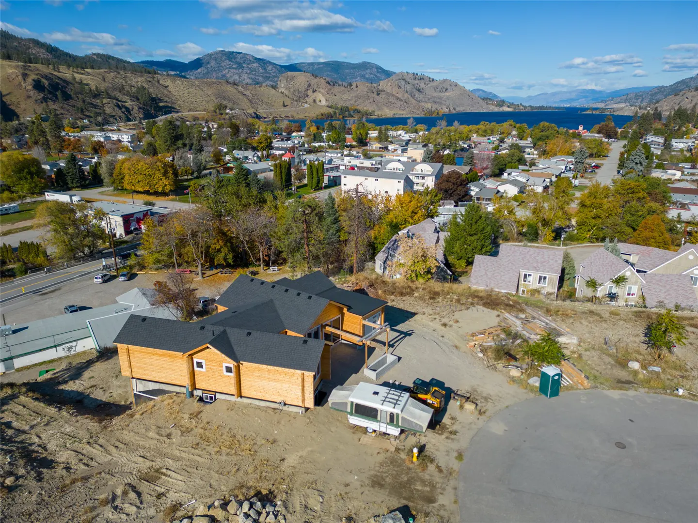Aerial view of a new, light-wood home with a dark roof, set against a backdrop of mountains, a lake, and a small town.