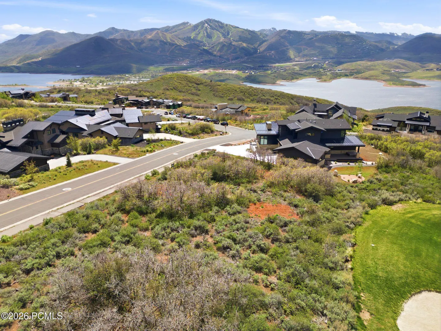 Aerial view of modern homes with dark roofs, a road, and a lake with mountains in the background on a sunny day.