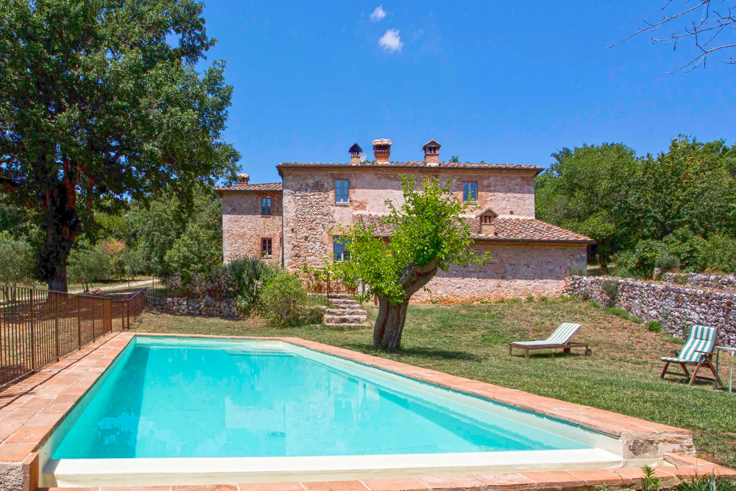Exterior view of a stone house with a pool, lawn chairs, and trees under a blue sky.