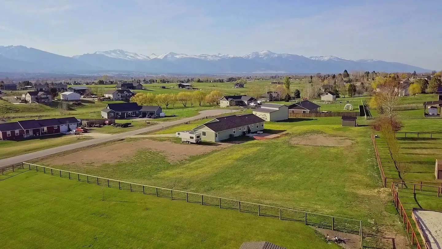 Aerial view of a rural neighborhood with green lawns, houses, fences, and snow-capped mountains in the background.