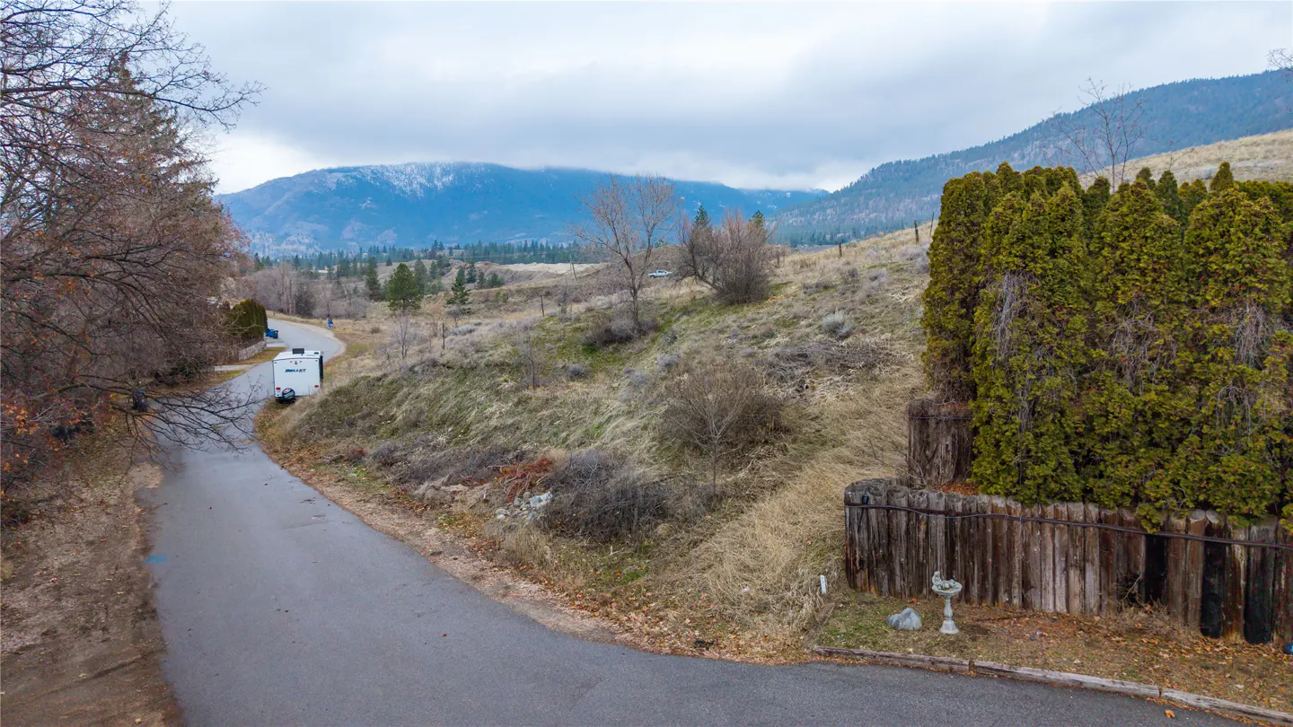 An elevated view of a vacant lot with a winding road, trees, and mountains in the background on a cloudy day.