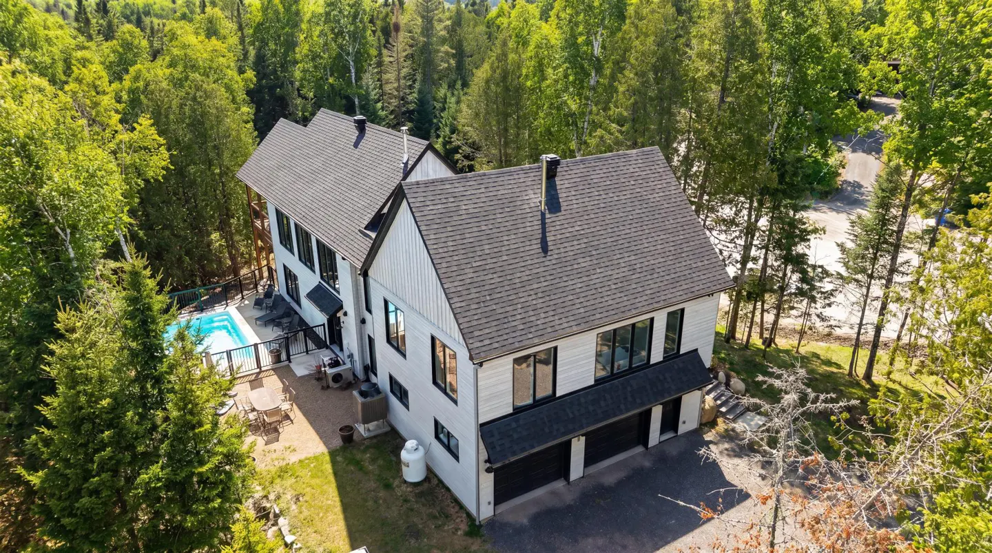 Aerial view of a modern white house with a dark gray roof, black trim, a pool, and a two-car garage surrounded by green trees.
