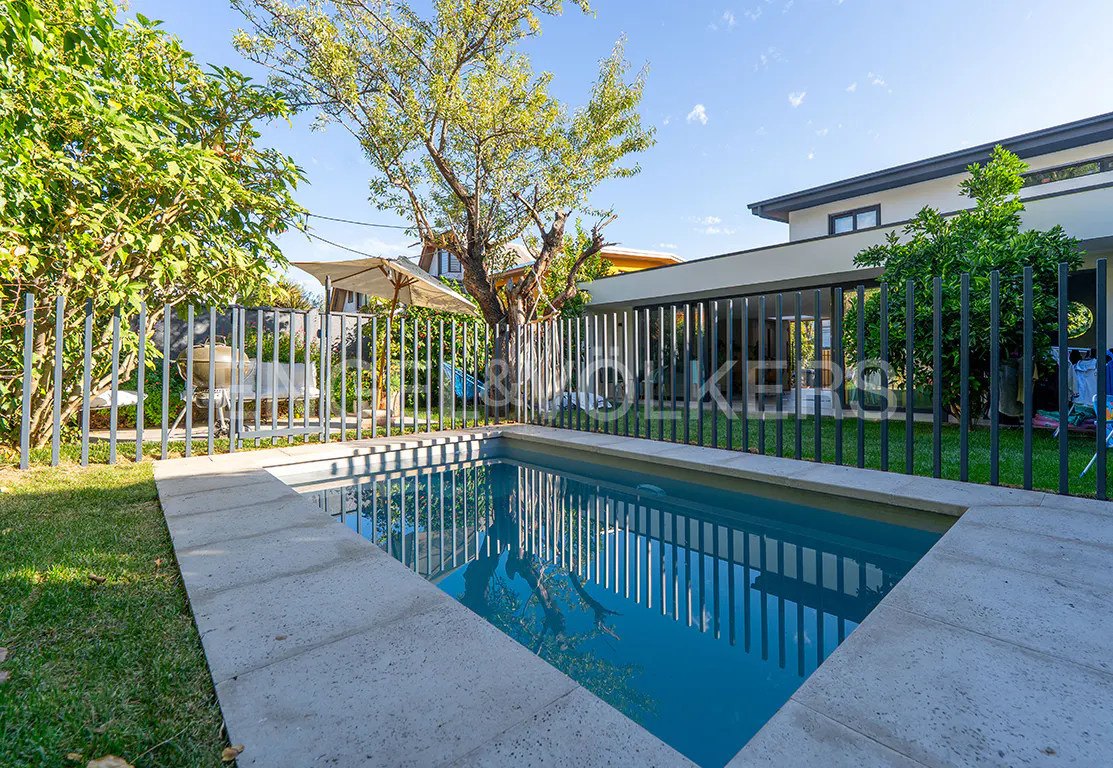 A rectangular pool with blue water is surrounded by a stone border and a black metal fence. Green grass and trees are in the background.