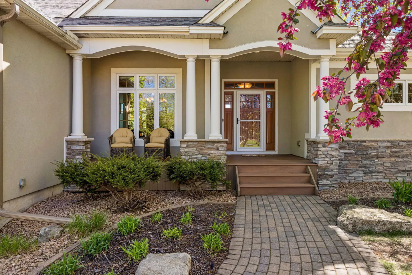 Front exterior of a beige stucco home with stone accents, white columns, and a brick walkway. Two chairs sit on the porch.
