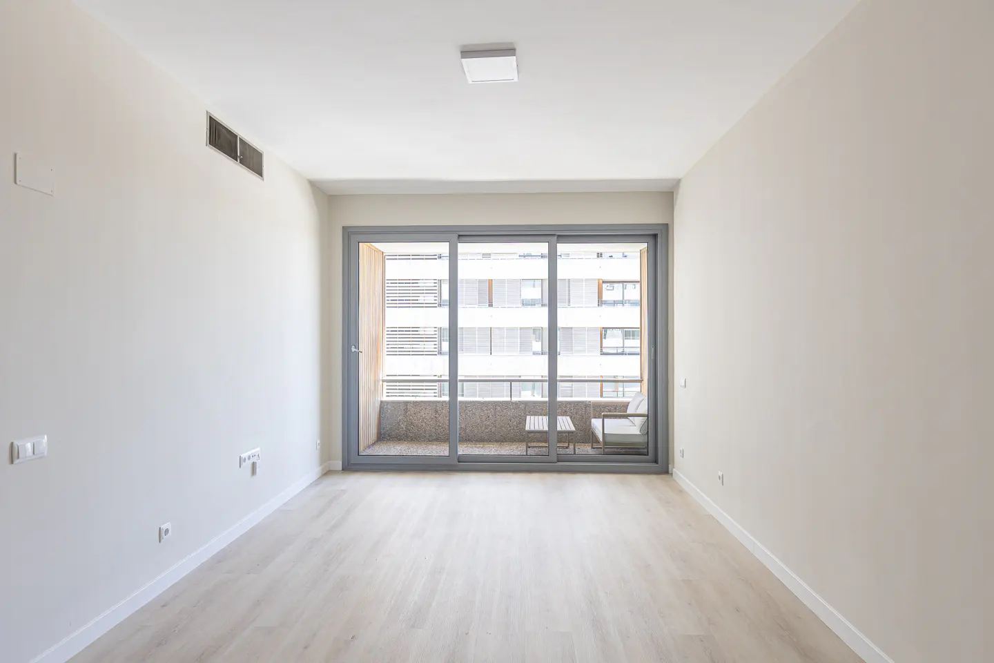 Empty room with light wood floors, white walls, and a large sliding glass door leading to a balcony with outdoor seating.
