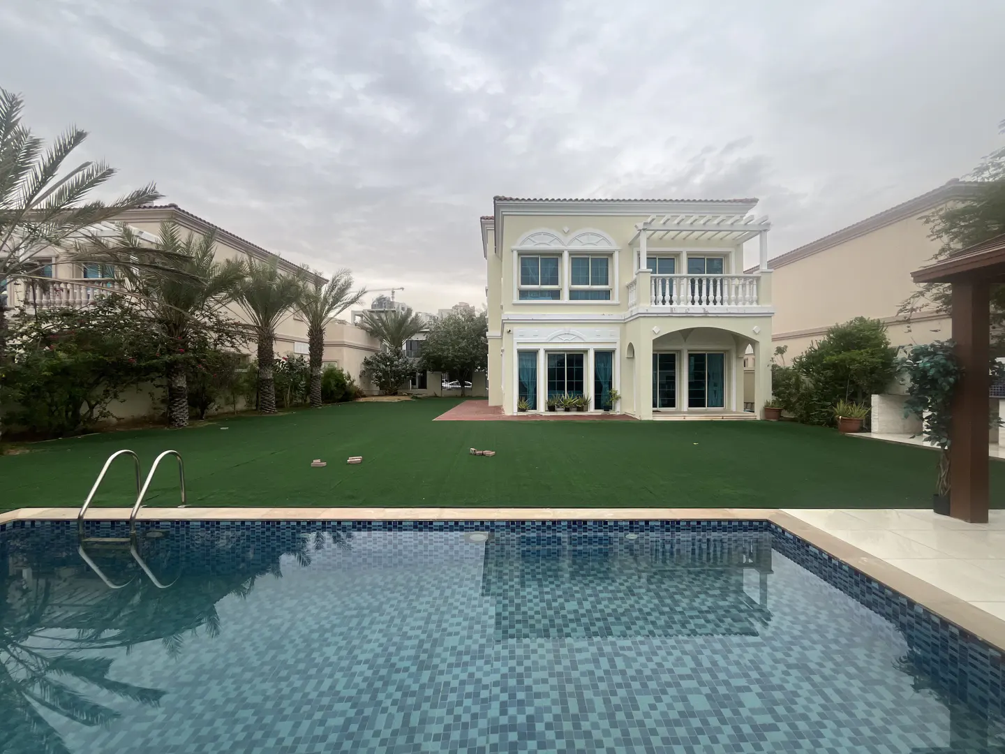 Backyard view of a two-story house with a pool, green lawn, and palm trees under a cloudy sky.