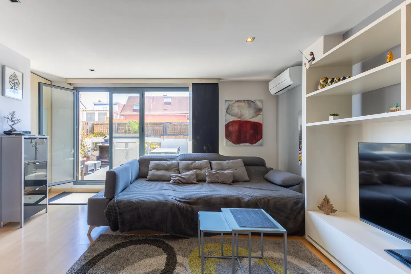 Bright living room with a gray sofa, pillows, and a rug. Sliding glass doors lead to a patio. White shelves and a TV are on the right.