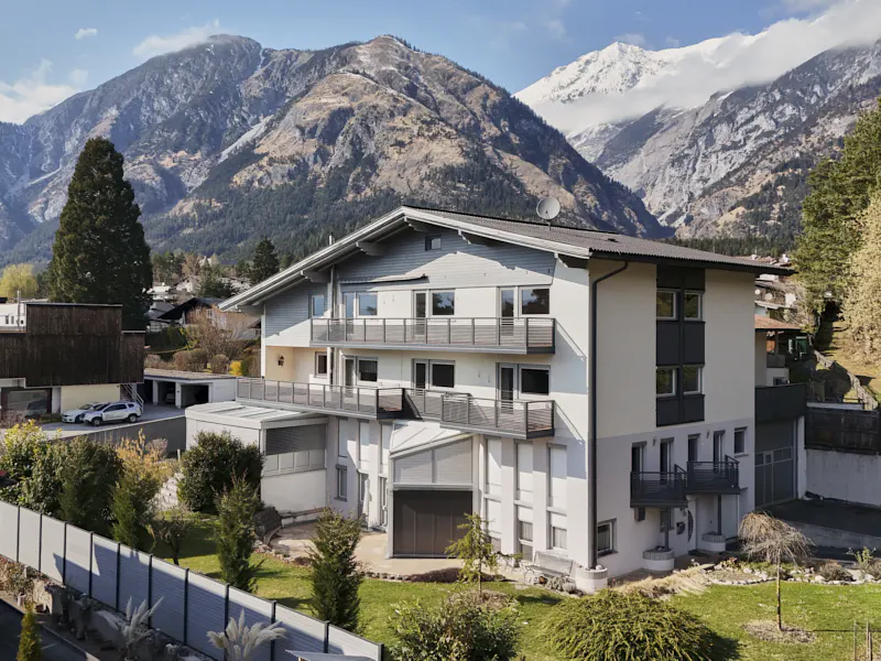 Three-story white house with balconies, set against a backdrop of mountains and trees.