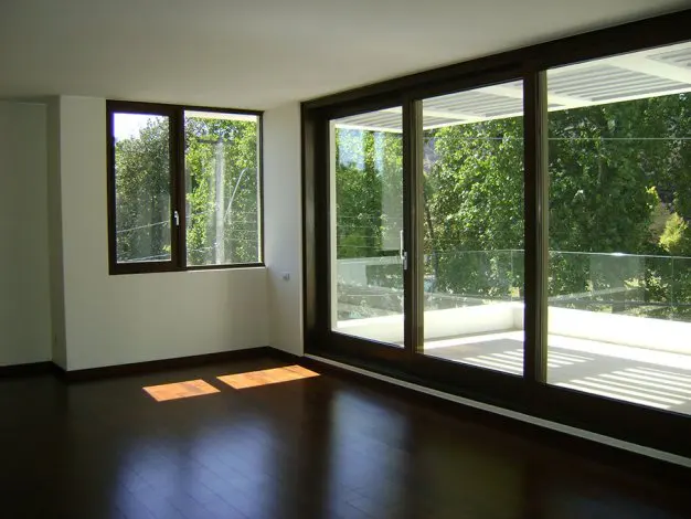 An empty room with dark wood floors, white walls, and large windows with a view of green trees.