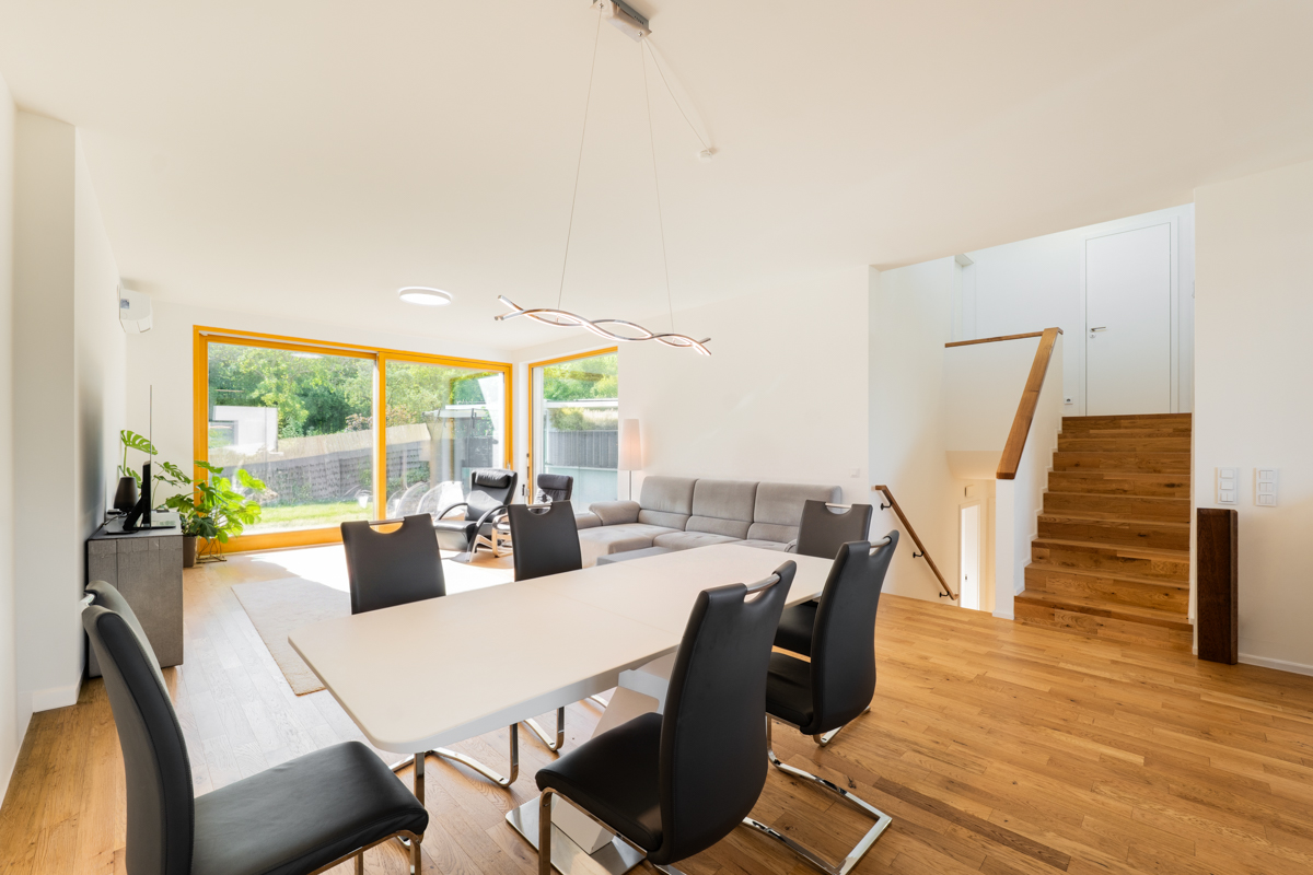 Modern dining area with white table, black chairs, and wooden floors, leading to a living room and wooden staircase. [AI-generated text]