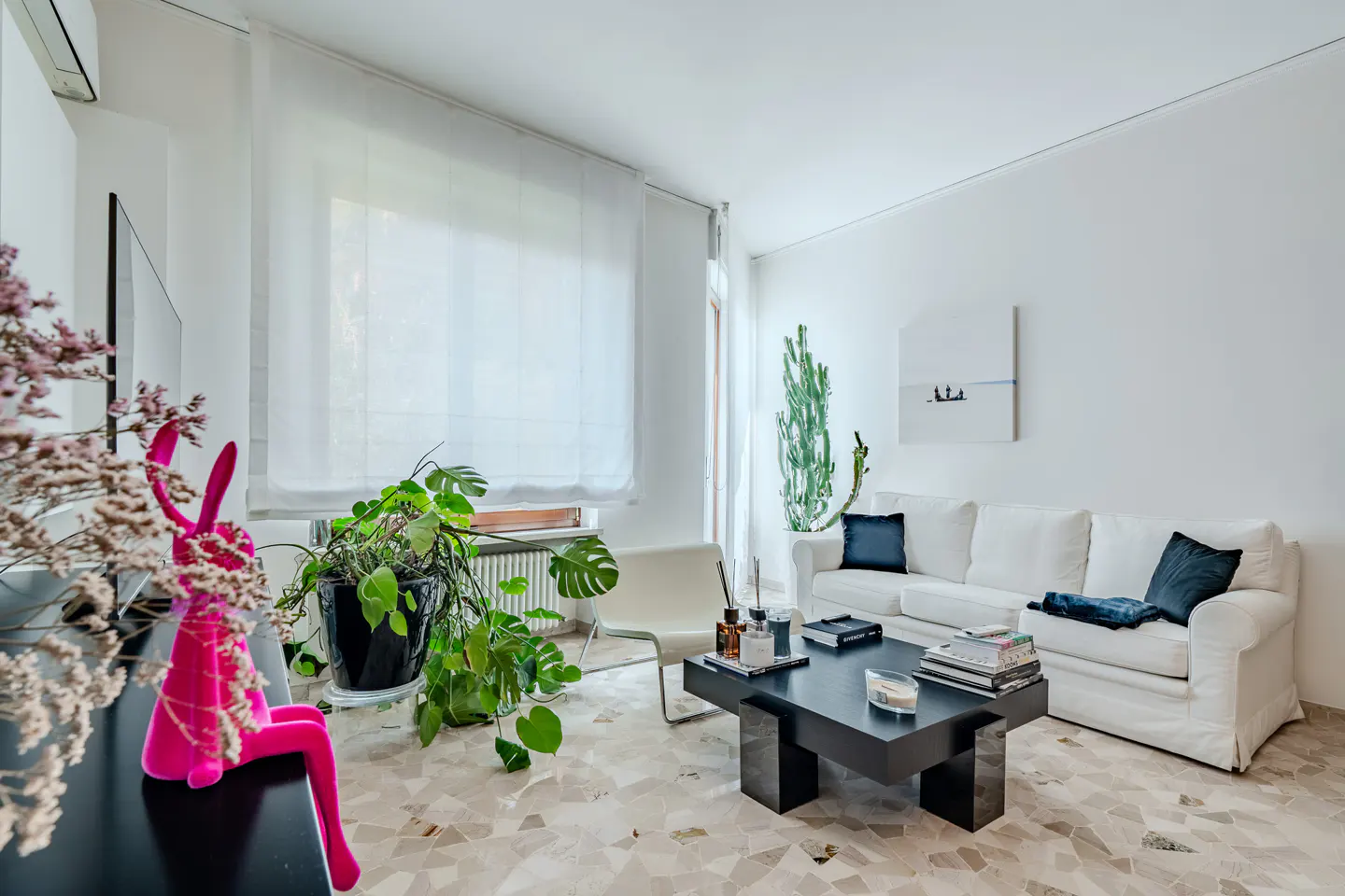 Bright living room with white walls, a white sofa with blue pillows, and a black coffee table on a patterned floor. A pink bunny statue sits on a black console.
