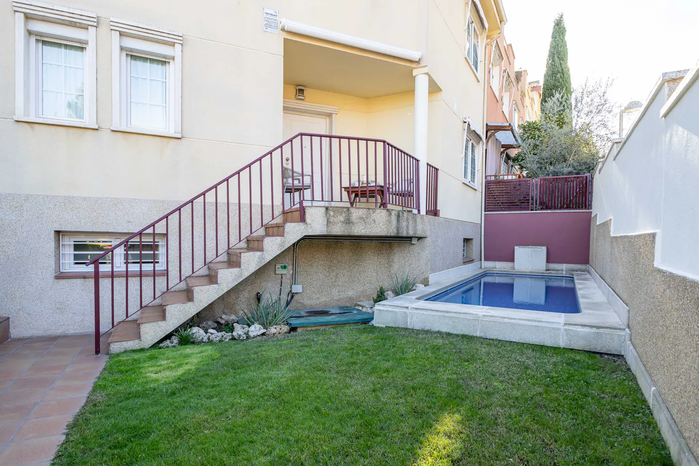 Backyard of a cream-colored house featuring a small rectangular pool, a green lawn, and stairs leading to a porch.