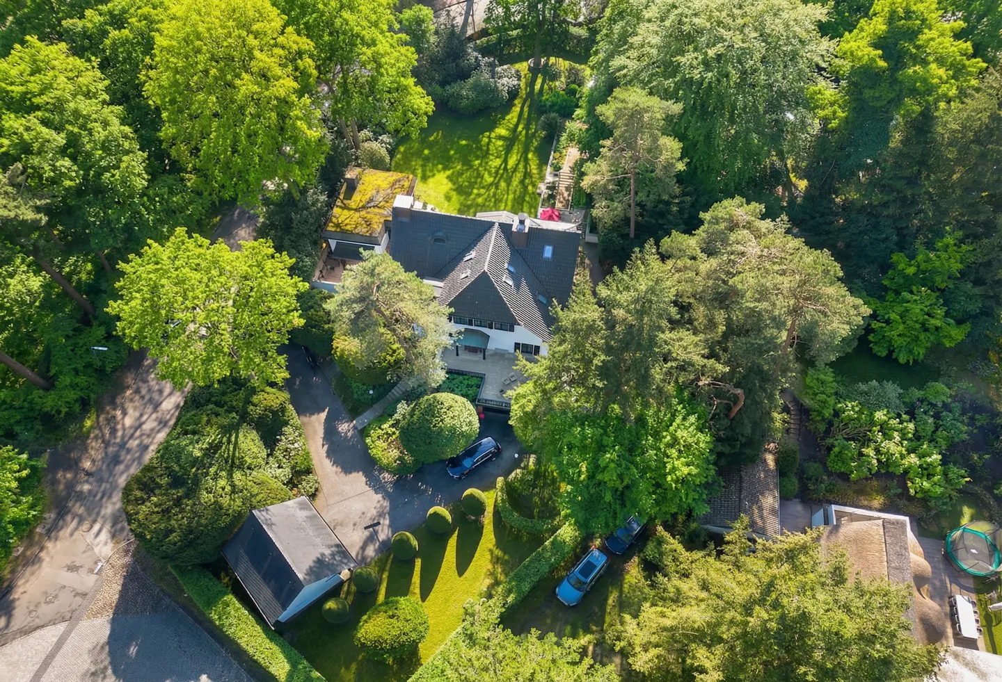 Aerial view of a white house with a dark roof, surrounded by green trees and manicured lawns. Two blue cars are parked in the driveway.