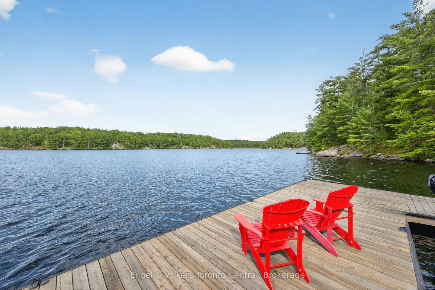 Two red Adirondack chairs sit on a wooden dock overlooking a calm lake surrounded by green trees under a blue sky.