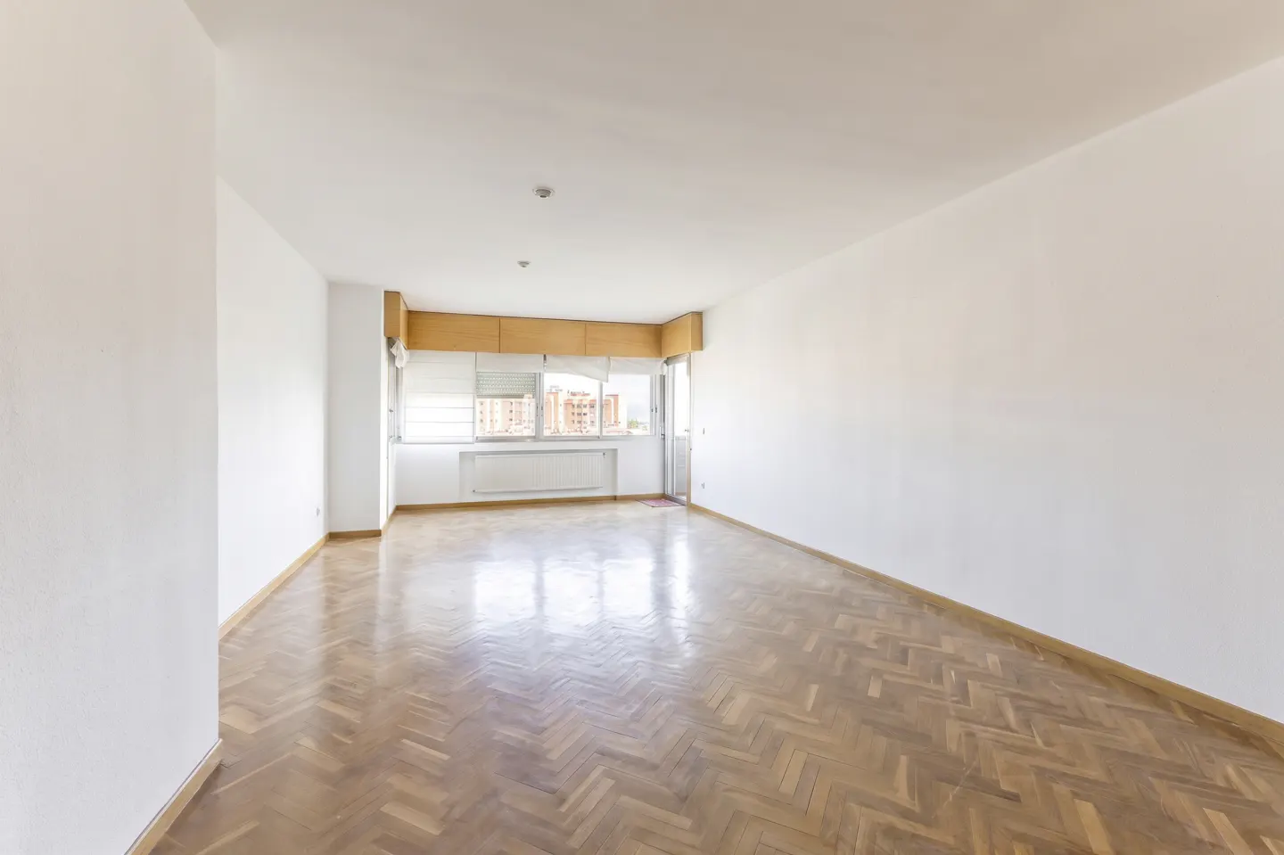 Bright, empty room with white walls and herringbone wood floor. A window with blinds and a door lead to a balcony.