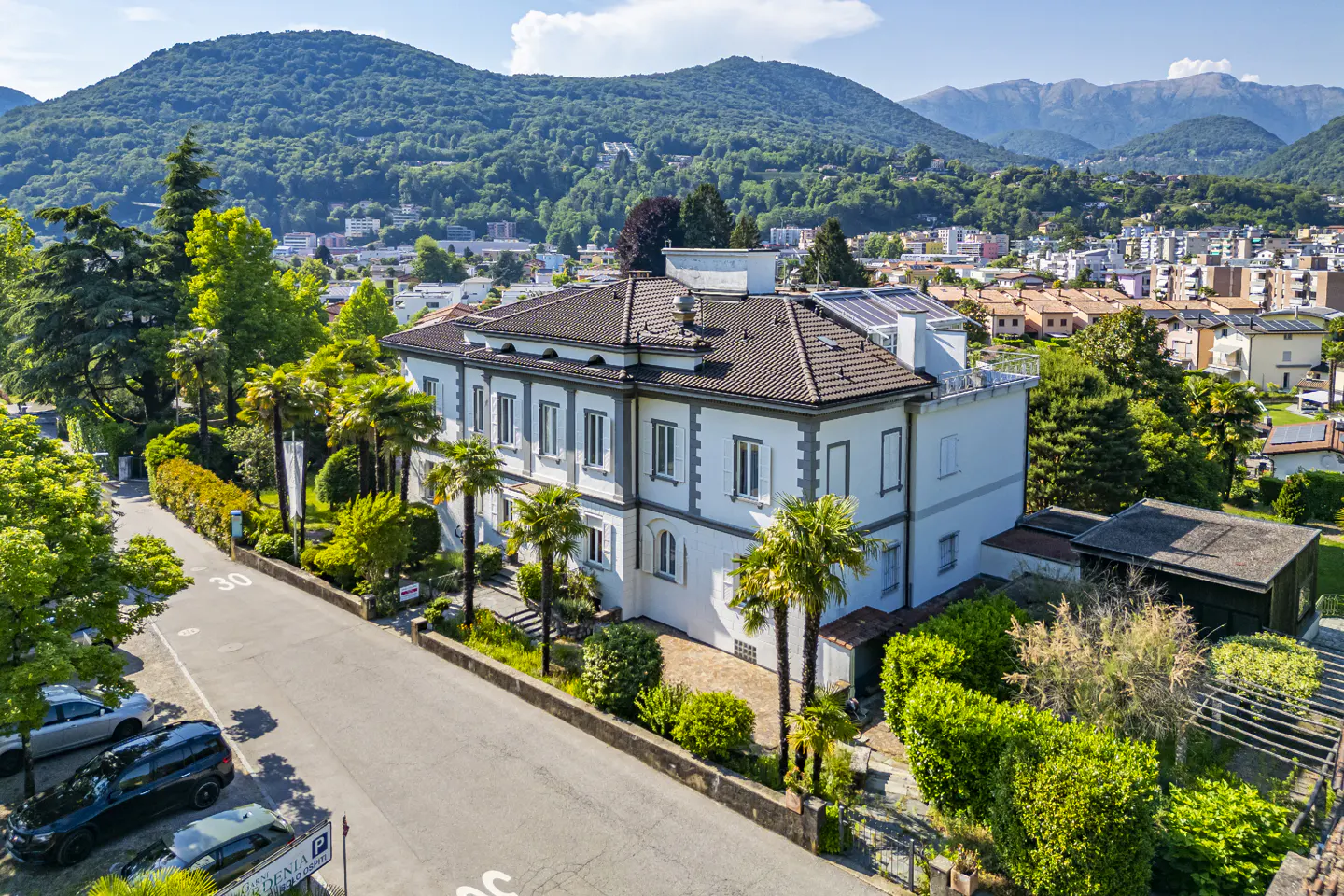 Aerial view of a white villa with a brown roof, surrounded by palm trees and lush greenery, set against mountain scenery.