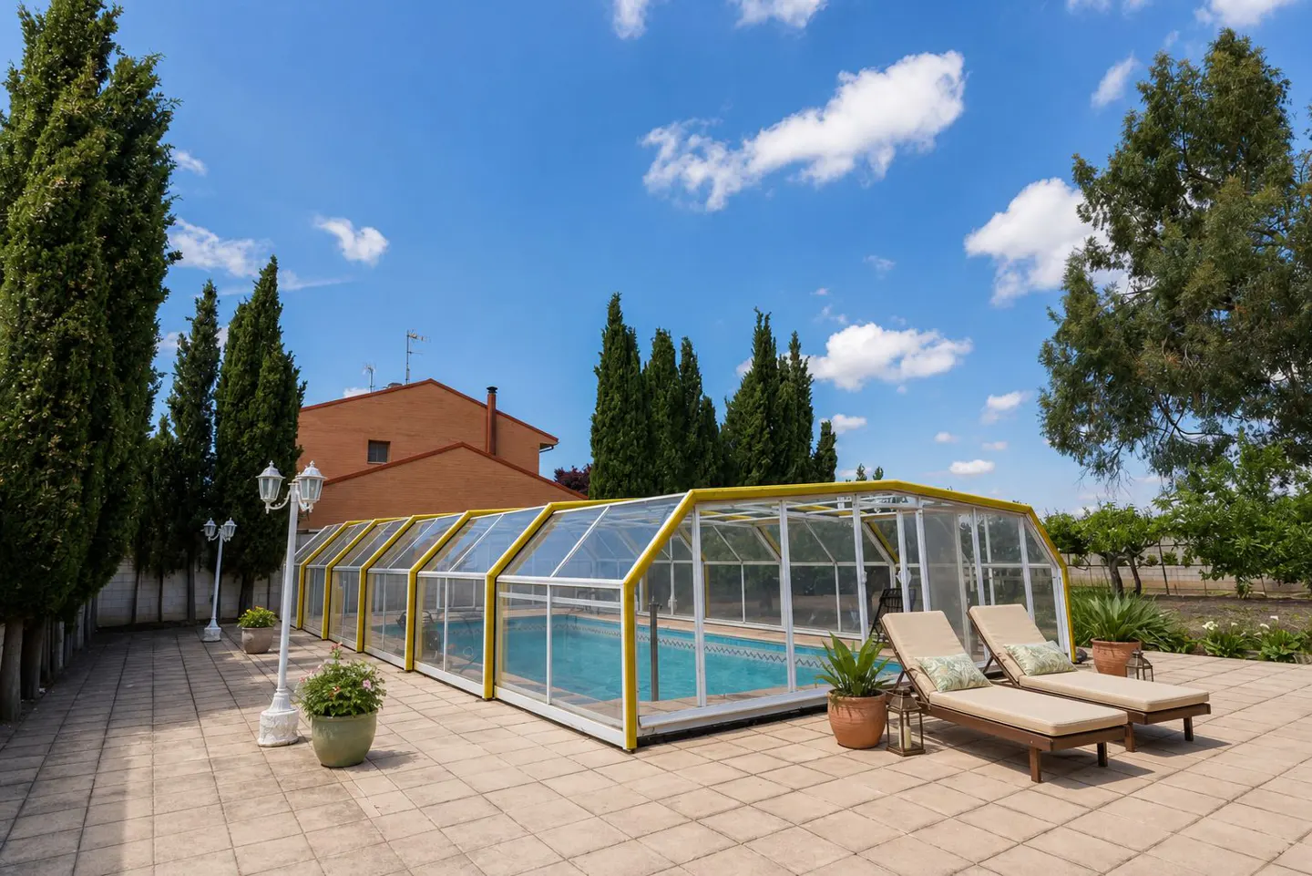 Outdoor pool with a yellow frame and clear enclosure, two lounge chairs, potted plants, and a blue sky with white clouds.
