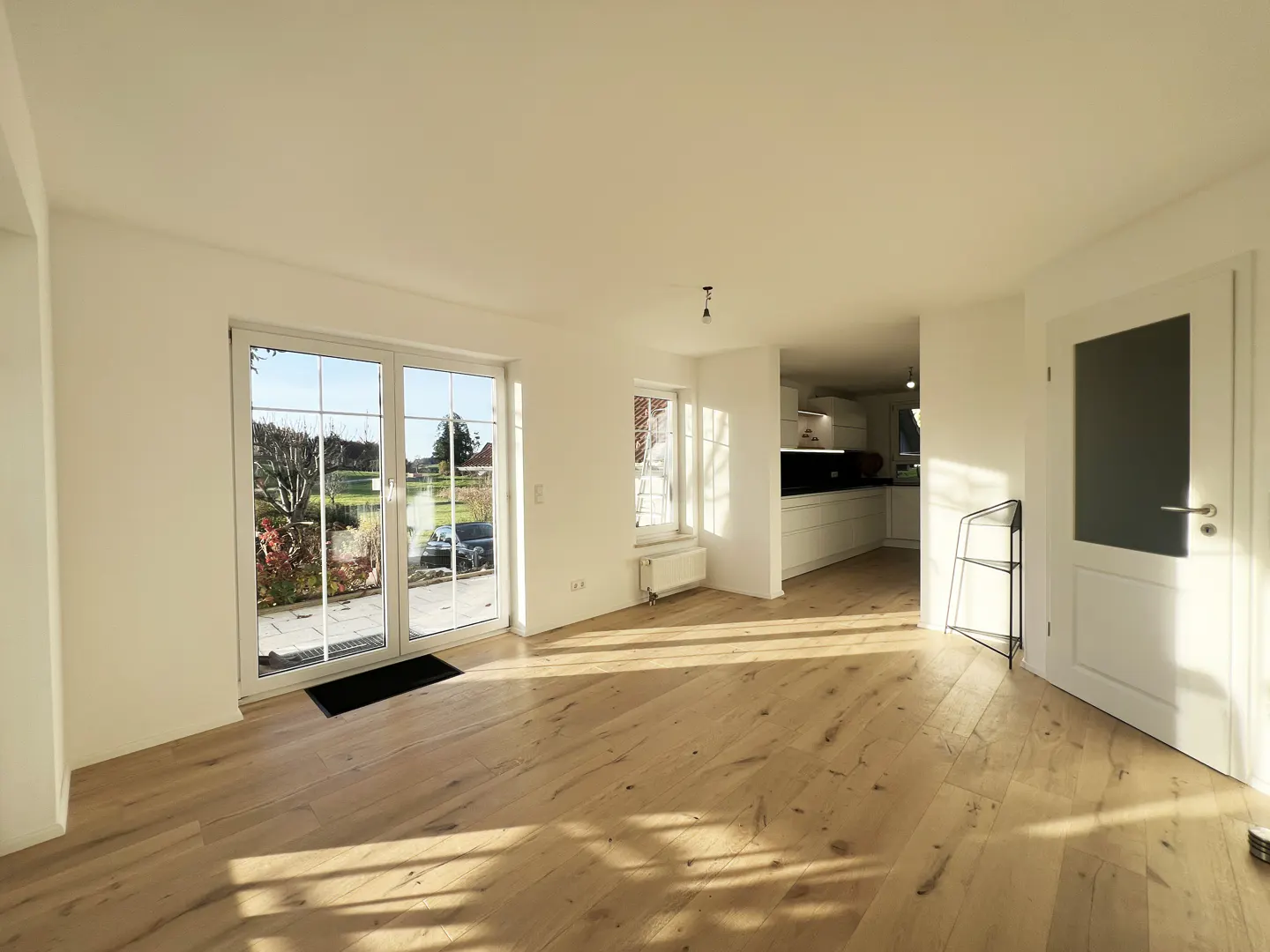 Bright, empty room with wood floors, white walls, and a view to the outside through glass doors. A kitchen is visible in the background.