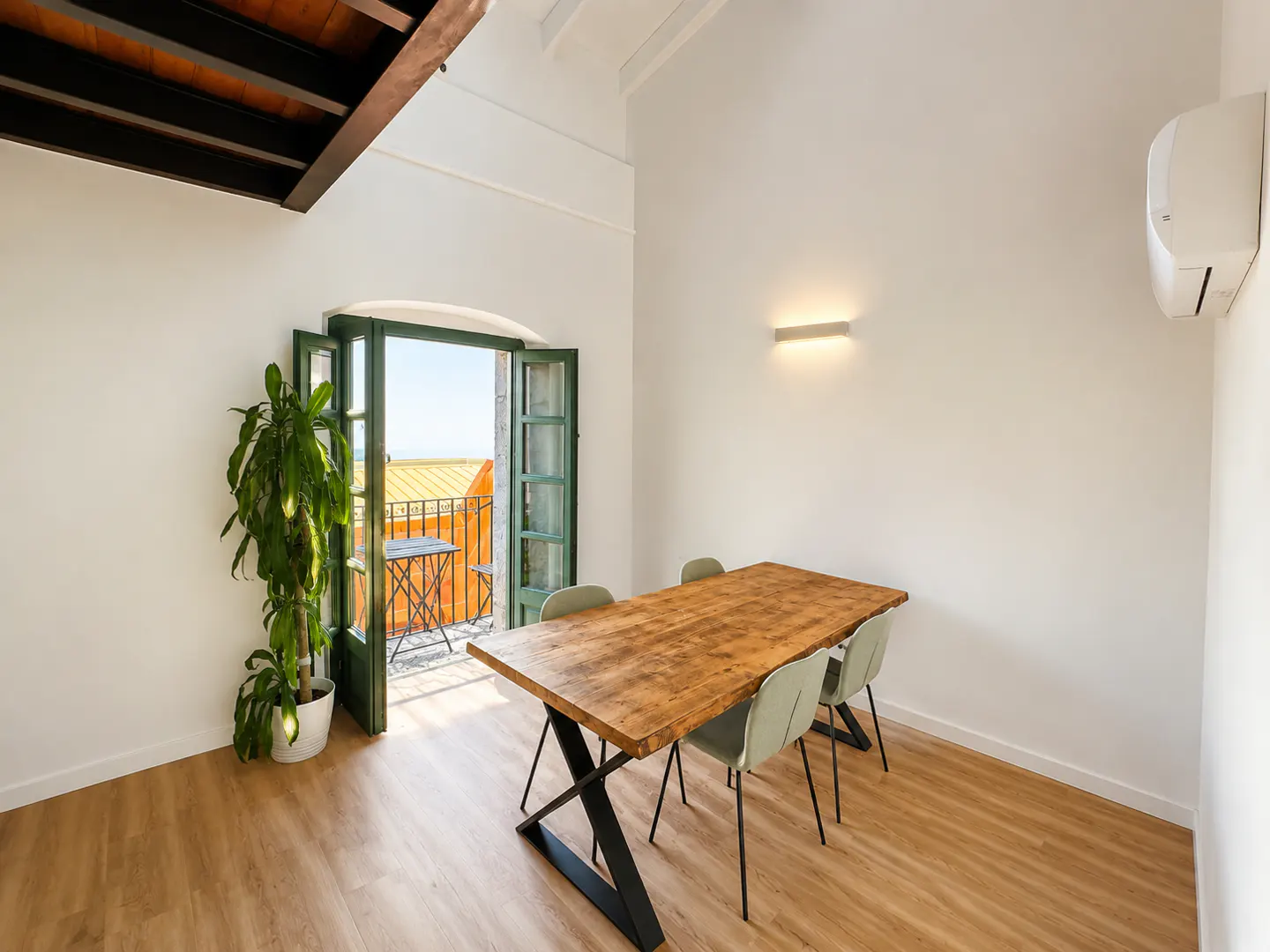 Bright dining room with wood table, four chairs, and open green doors to a balcony with a table and orange buildings.