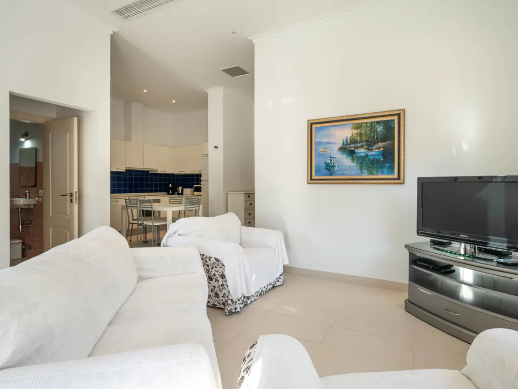 Bright living room with white sofas, a TV, and a framed painting of boats. A kitchen and bathroom are visible in the background.