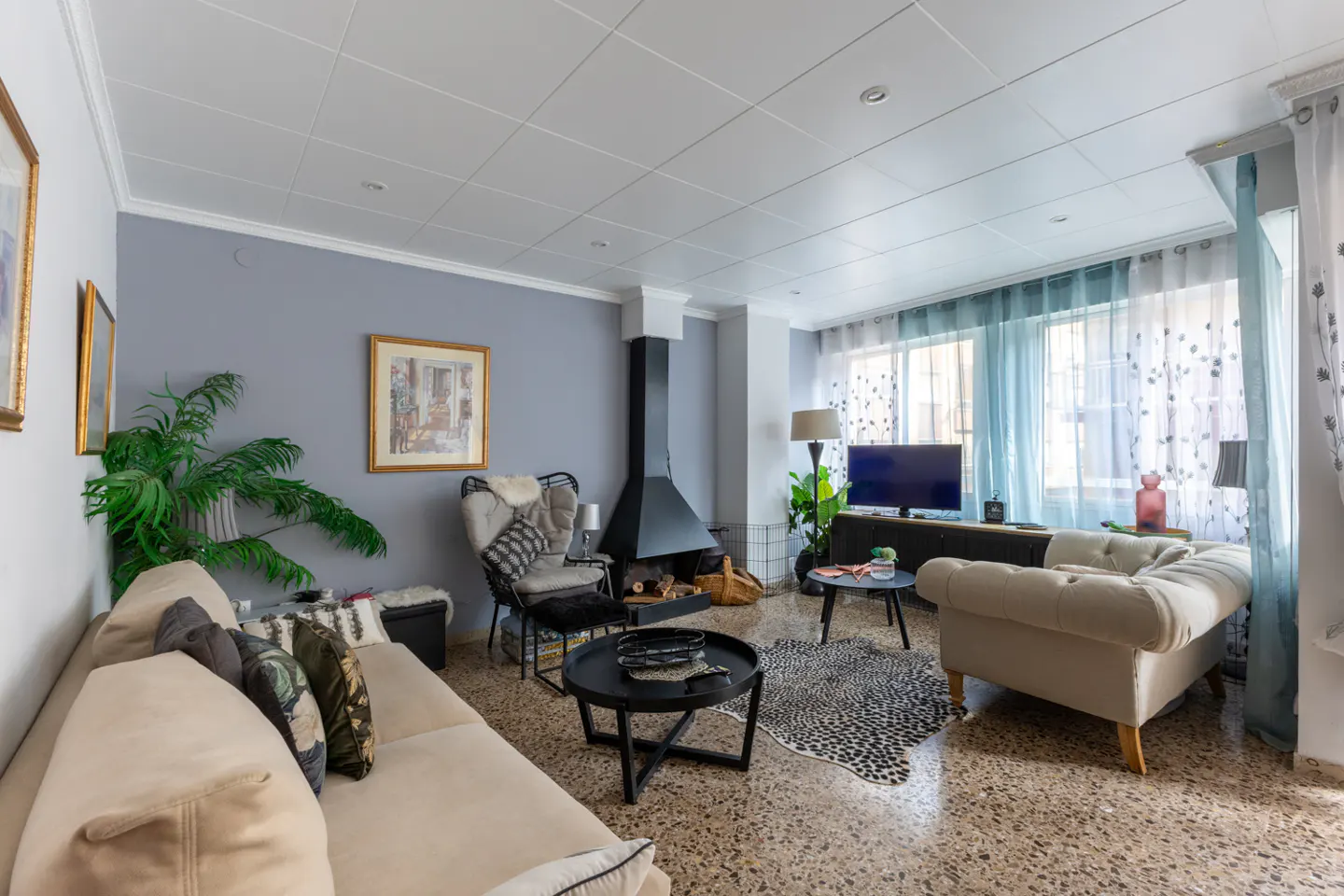 Living room with beige sofa, black fireplace, TV, and blue curtains. A patterned rug lies on the terrazzo floor.