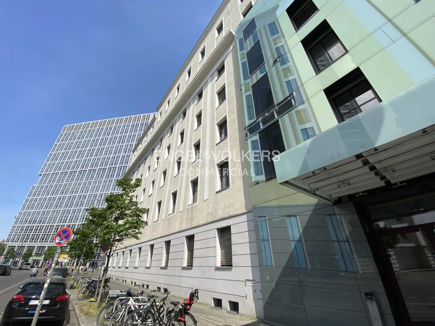 Exterior view of a modern commercial building with glass facade and a beige building next to it. Bicycles are parked on the sidewalk.
