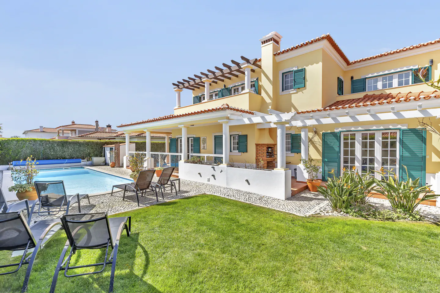 Backyard view of a two-story yellow house with a pool, green shutters, and a red tile roof. Lounge chairs sit on the lawn.