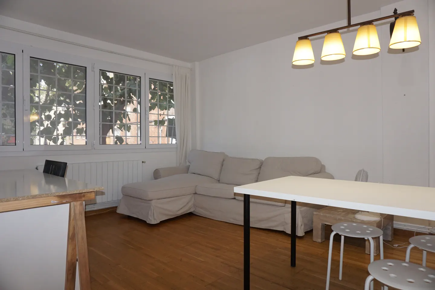 Bright living room with hardwood floors, a beige sectional sofa, a white table with stools, and a window with a view of trees.