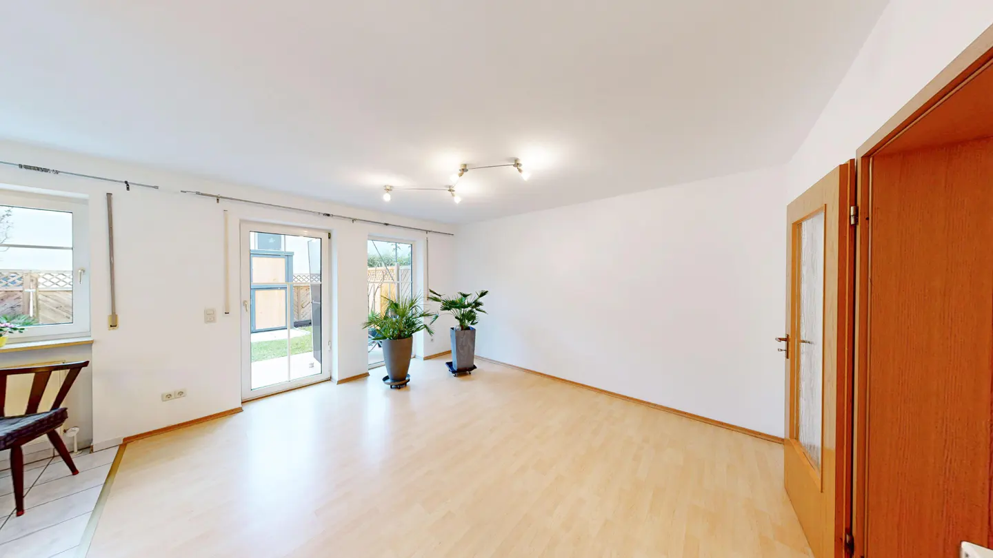 Bright, empty room with light wood floors, white walls, and two potted plants. Windows and glass doors lead to a garden.