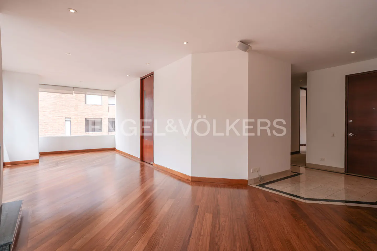 Empty room with polished hardwood floors, white walls, recessed lighting, and dark wood doors leading to a hallway.