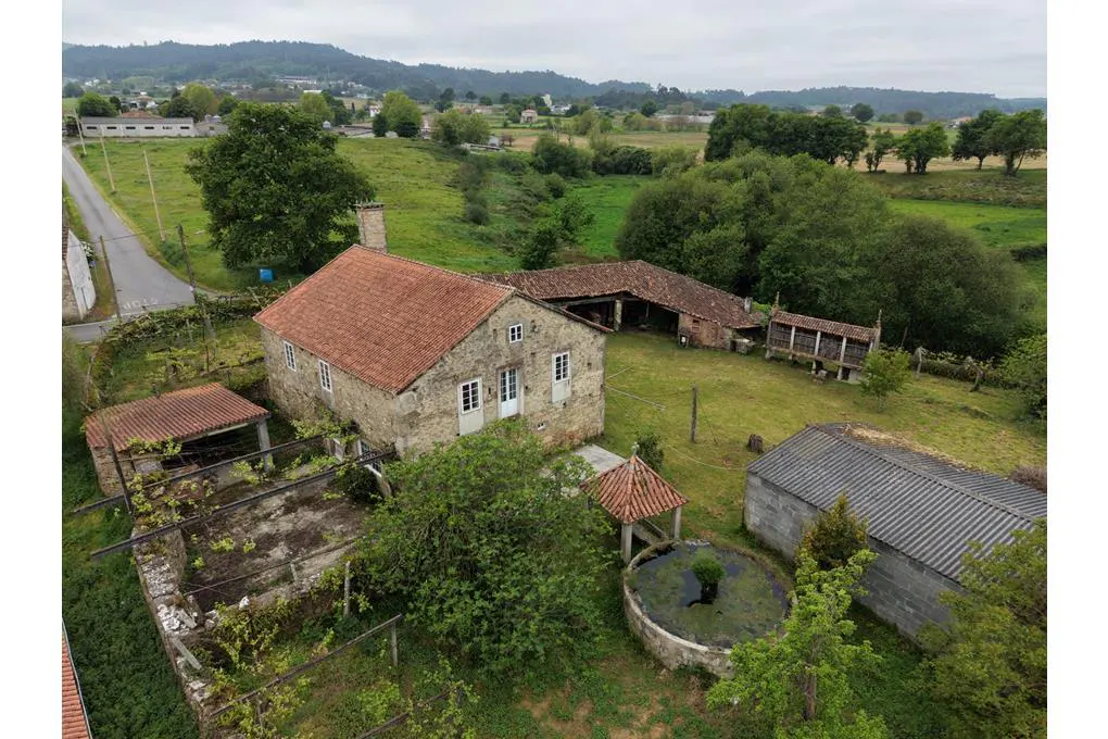 Aerial view of a stone farmhouse with a red tile roof, outbuildings, a small pond, and lush green rural surroundings.