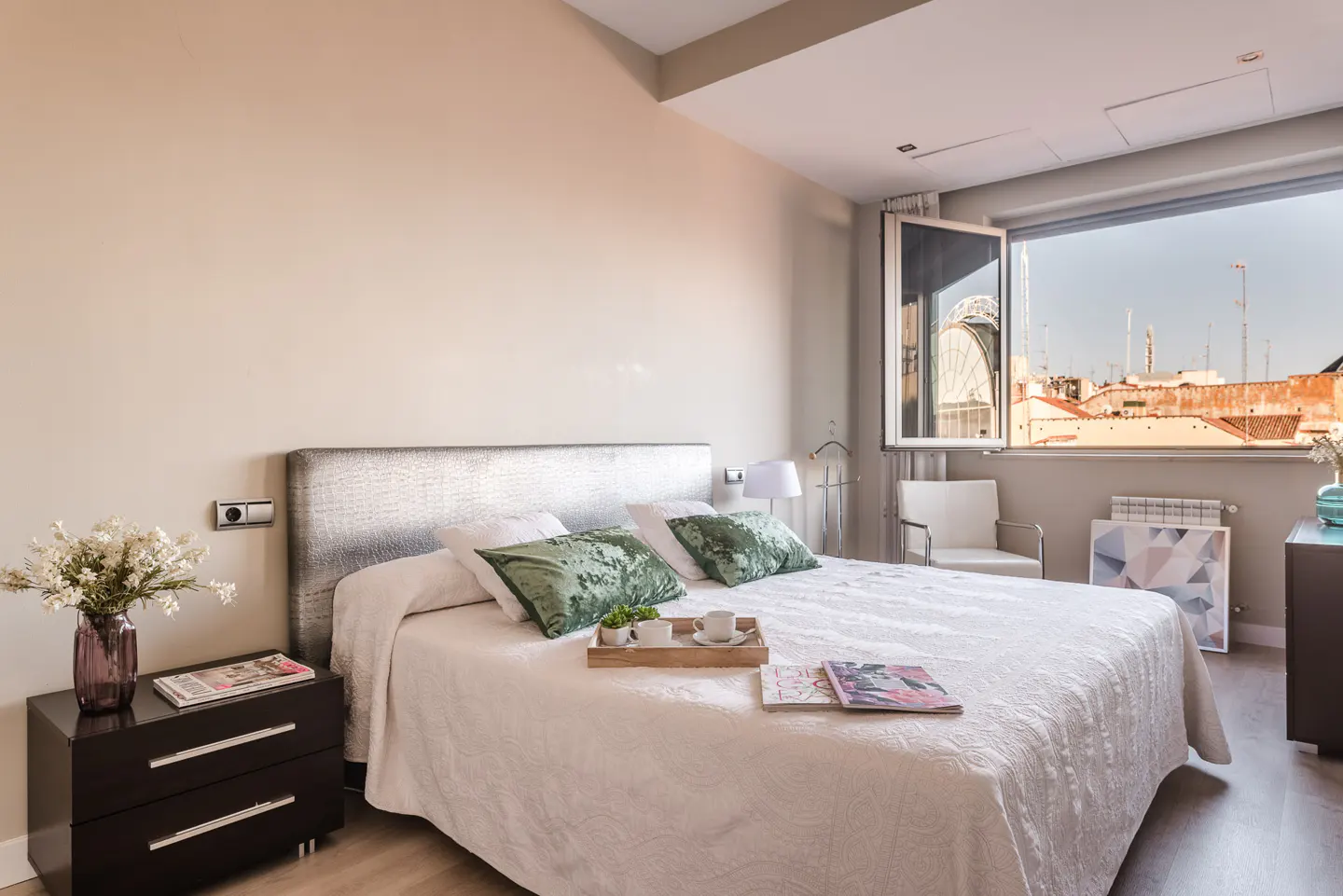 A bright bedroom with a large window showing a city view. A bed with white linens and green pillows is centered. A dark nightstand with flowers sits beside it.