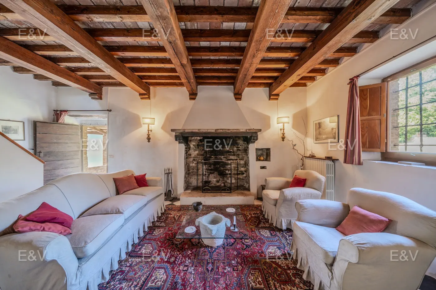 Living room with exposed wood beams, fireplace, white sofas, and a red patterned rug.