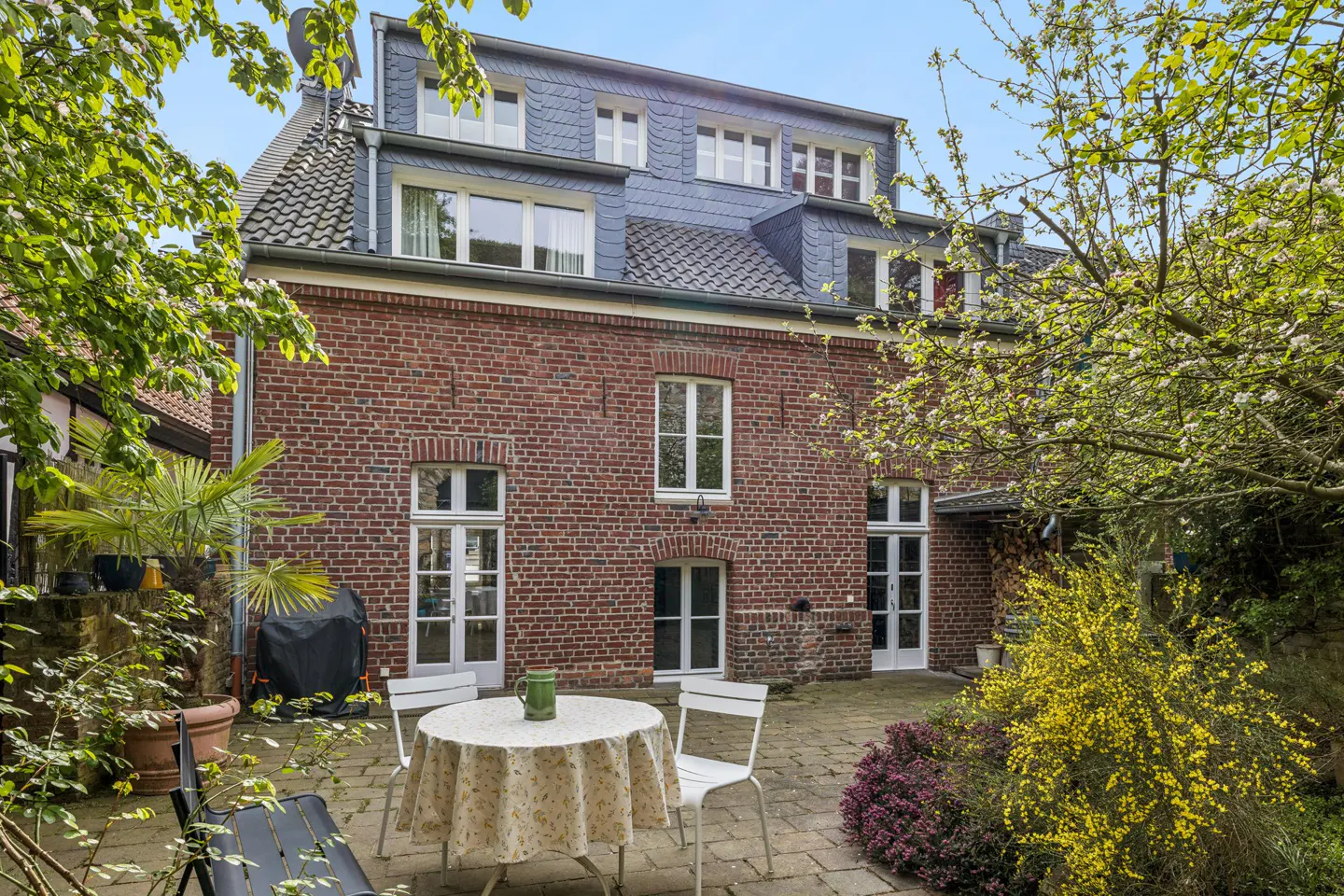 Brick house with a slate roof and white windows, seen from a patio with a table, chairs, and greenery.