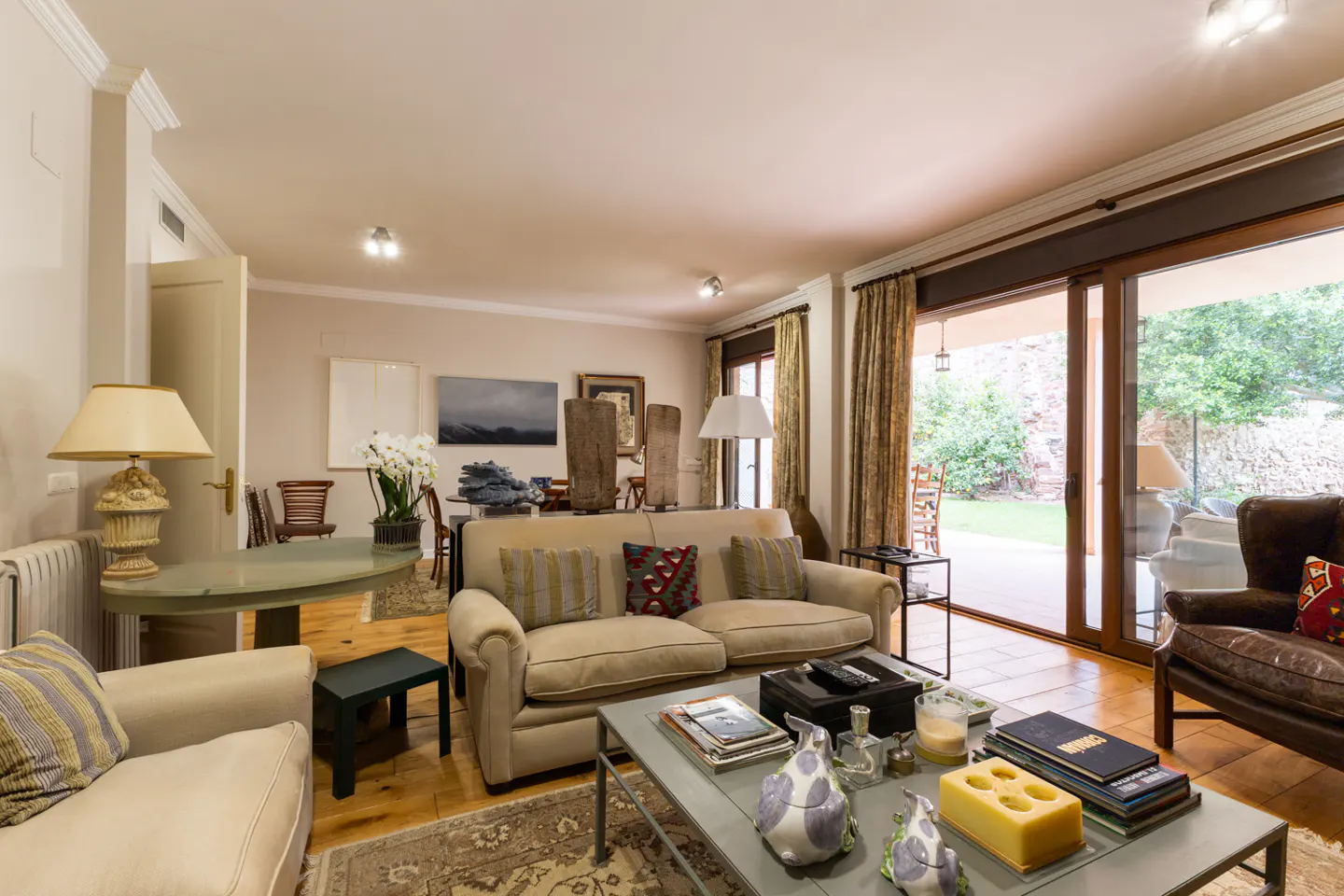 A bright living room with beige sofas, a gray coffee table, and a view of a green garden through sliding glass doors.