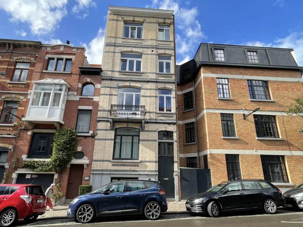 Street view of three brick buildings with parked cars. The center building is tall and narrow, with a small balcony.