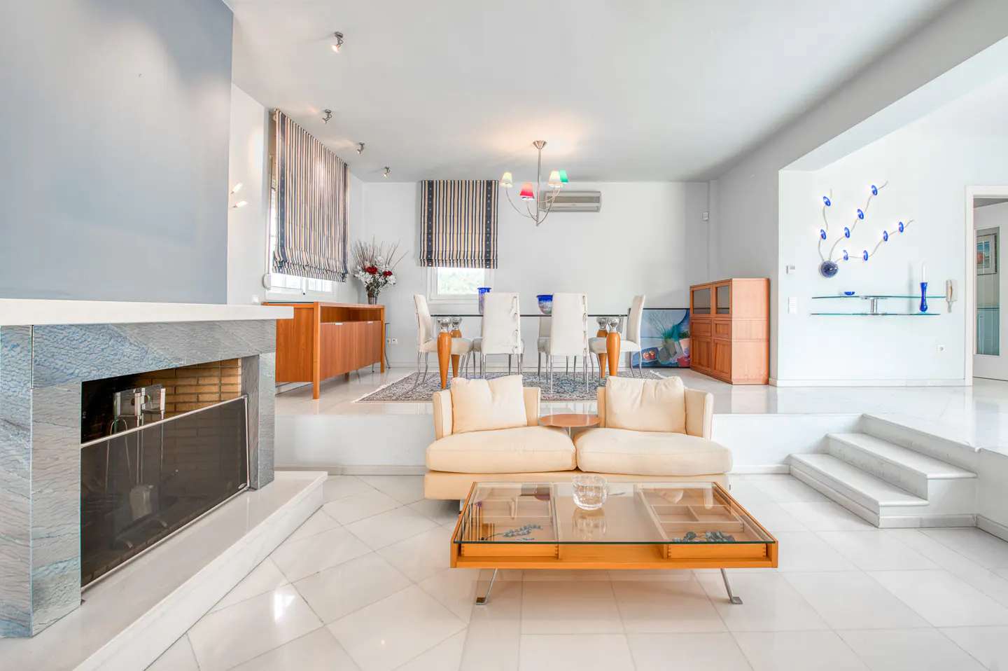 Bright, modern living room with a fireplace, cream sofas, and a glass-topped coffee table. A dining area is visible in the background.