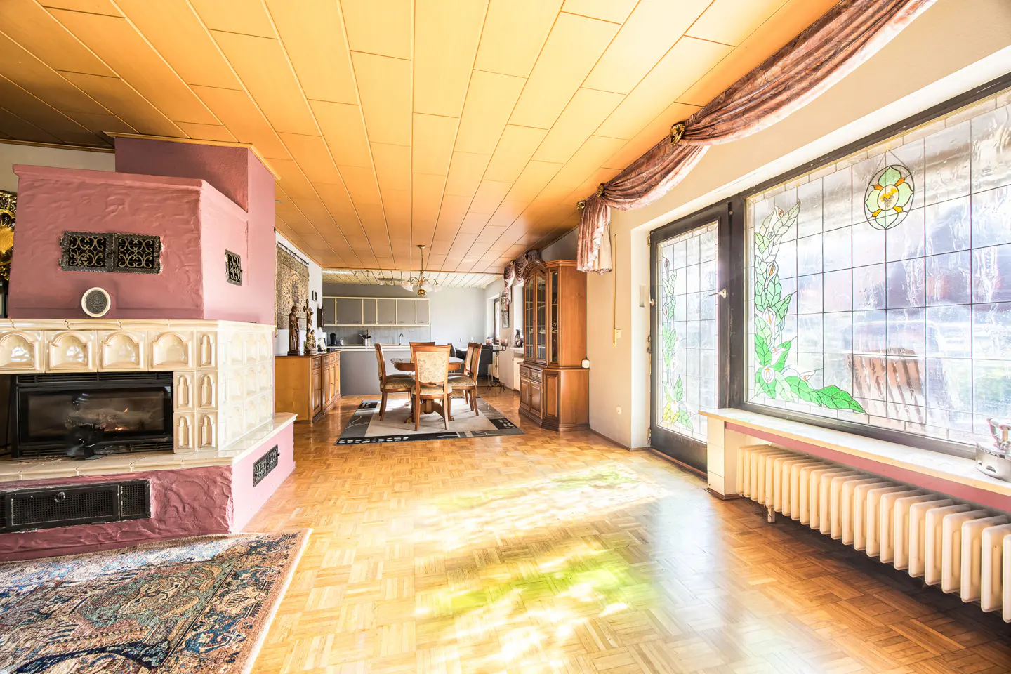 A bright living room with a pink fireplace, wood floors, and stained glass windows. A dining table and kitchen are visible in the background.