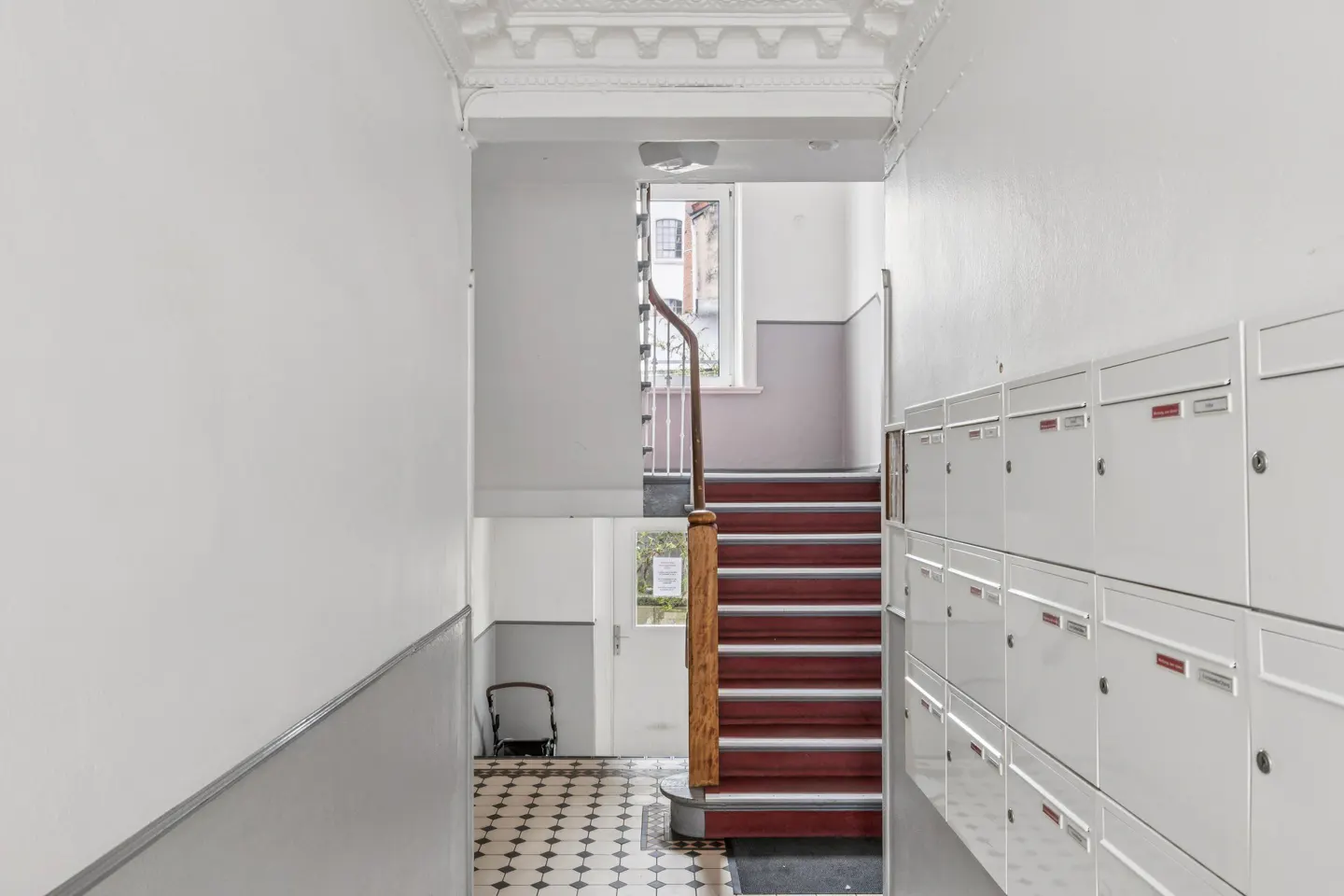 Hallway with white walls, mailboxes, and a staircase with red steps and a wooden banister leading to an upper level.