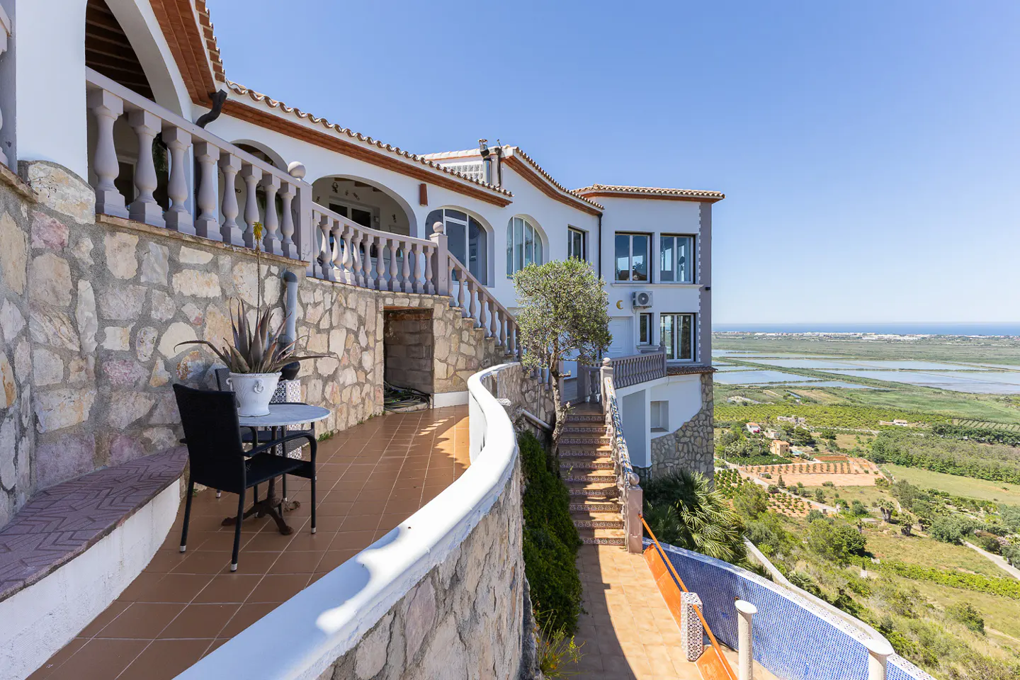 Exterior view of a white multi-story house with stone walls, a patio with a table and chairs, and a scenic view of the landscape.