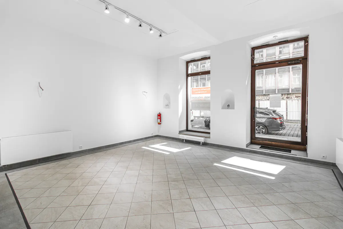 Bright, empty room with white walls, tile floor, and track lighting. Windows and a door show a street scene outside. A red fire extinguisher is visible.
