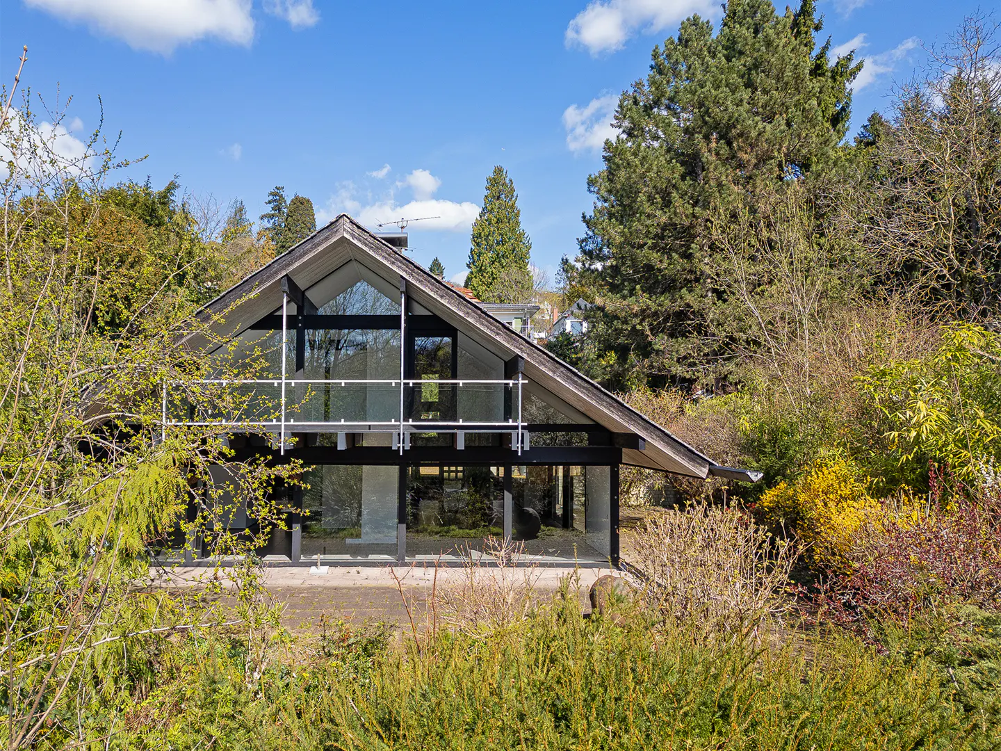 A modern A-frame house with large glass windows, surrounded by lush greenery and trees under a blue sky.