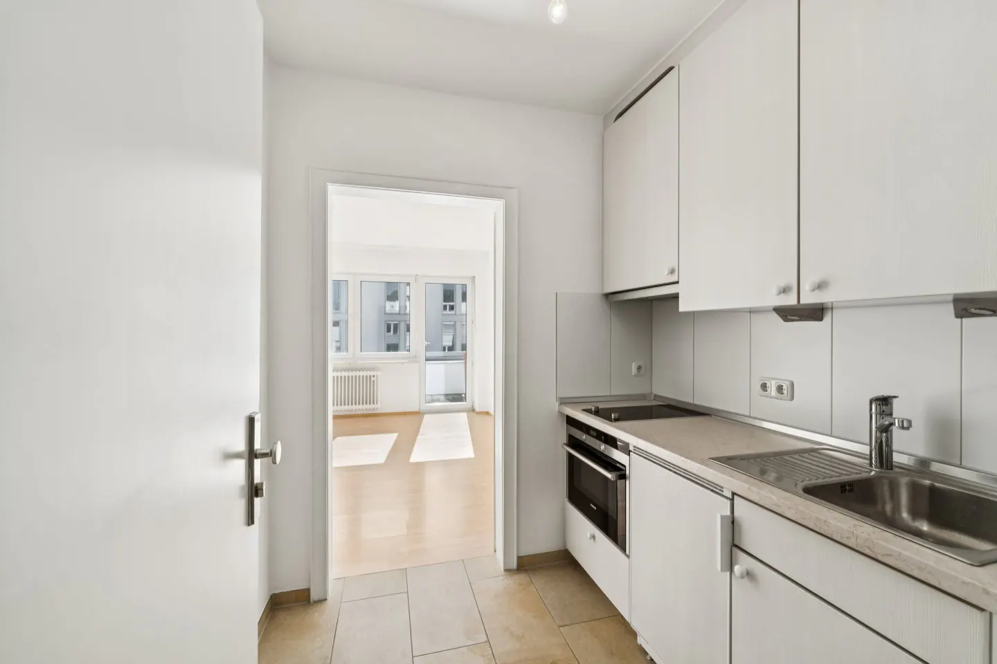 A bright, modern kitchen with white cabinets, a stainless steel sink, and a view into a sunlit living room.